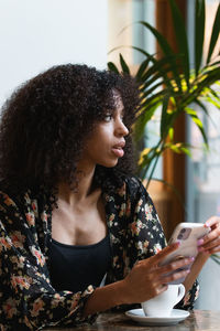 Young ethnic female browsing on cellphone while sitting looking away at table with a cup of coffee