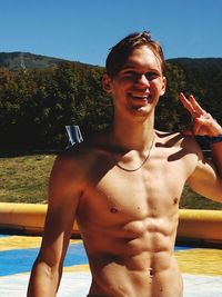 Portrait of shirtless young man in swimming pool showing peace sign on sunny day