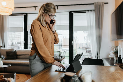 Side view of businesswoman talking on phone while using laptop at home