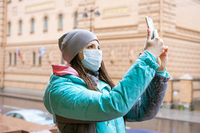 Young woman in a protective mask in a jacket takes pictures on the phone