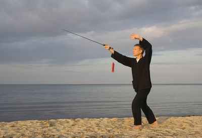 Mature man practicing tai chi with sword at beach against cloudy sky