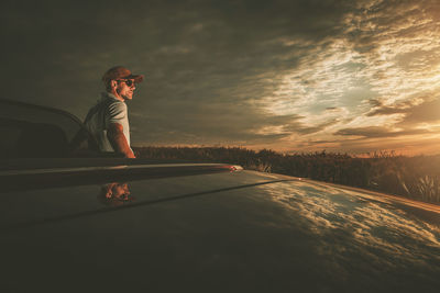 Man wearing sunglasses standing by car against sky during sunset