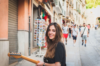 Portrait of young woman playing guitar while standing on sidewalk in city