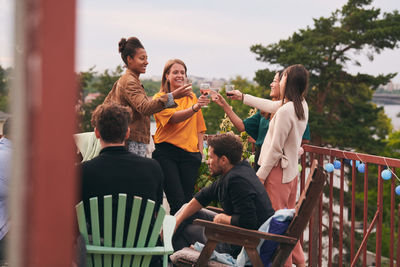 Female friends toasting drinks while men sitting on chairs during party