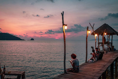 People sitting on pier over sea against sky during sunset