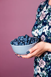 Midsection of woman holding blueberries in bowl against colored background