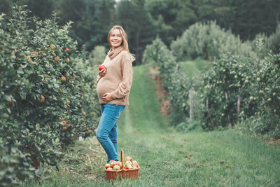 Smiling pregnant woman holding apple while standing by basket and trees