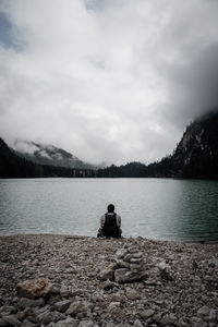 Rear view of man sitting on rock by lake against sky