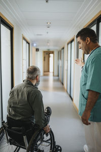 Male doctor showing way to male patient sitting in wheelchair at hospital corridor