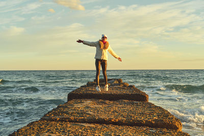 A woman with blond hair in a hat and a fur coat walks along the sea along a stone breakwater