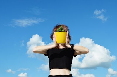 Woman covering face with potted plant against blue sky