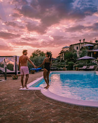 Couple standing by swimming pool against sky during sunset