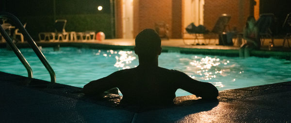 Rear view of silhouette man sitting by swimming pool at night