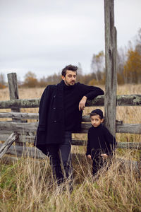 Father and son in black clothes stand in a field with an old wooden horse fence in the fall