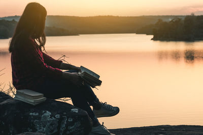 Side view of woman with book sitting by river during sunset