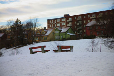 Empty bench on snow in front of house against sky