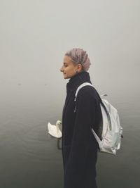 Side view of young woman with backpack standing by lake during foggy weather