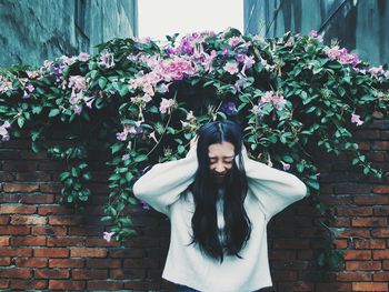 Woman covering ears while standing against plants and brick wall in yard