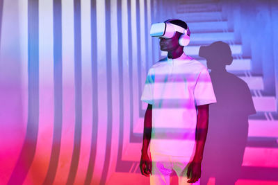 Young black man in white t shirt with modern vr headset in studio with bright colorful neon illumination