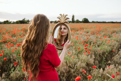 Beautiful young woman by poppy flowers on field looking at her reflection