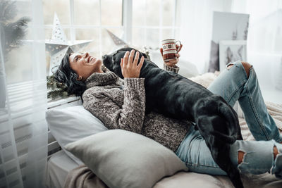 Laughing woman holding coffee cup playing with dog on bed