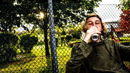Young man drinking beer with eyes closed against chainlink fence at park