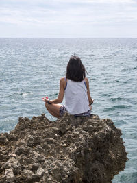 Rear view of woman meditation on rock against sea