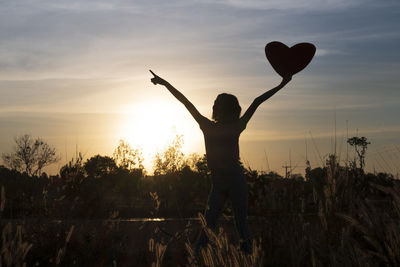 Silhouette woman standing by heart shape against sky during sunset