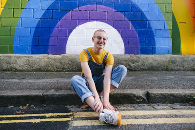Happy transgender person sitting on footpath in front of rainbow wall