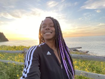 Portrait of smiling young woman standing by sea against sunset sky