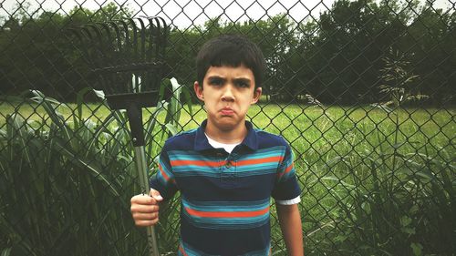 Portrait of sad boy holding gardening equipment while standing against plants and fence