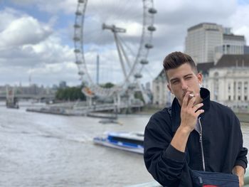 Young man smoking cigarette while standing against river in city