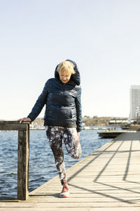 Senior woman stretching leg while standing on pier against clear sky