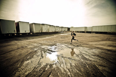 Athlete jogging on muddy field by cargo containers against sky