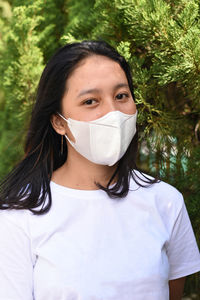 Portrait of young woman wearing mask standing behind tree outdoors