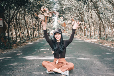 Portrait of smiling young woman throwing leaves while sitting on road amidst trees in forest