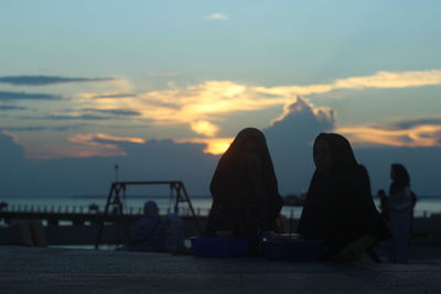 Silhouette female friends sitting at beach against sky during sunset