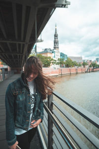 Smiling young woman with tousled hair standing on footbridge by river in city