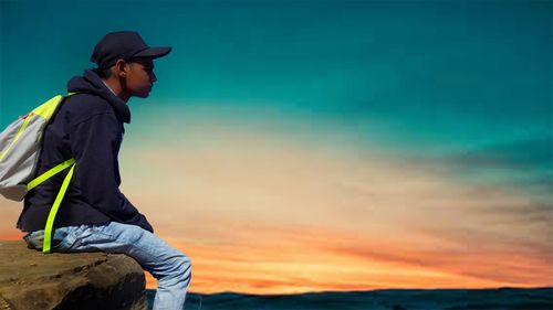 Side view of young man sitting on rock against sky during sunset