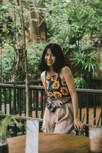 Portrait of smiling young woman standing by railing against plants