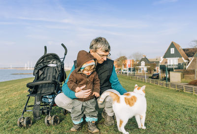 Grandfather with granddaughter looking at cat standing on grassy field against sky