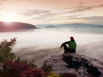 Man tourist sit on rock empire. view point with heather and branches above mist. sunny daybreak