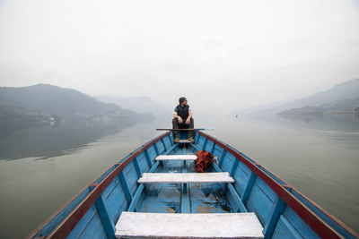 Male explorer floating in blue wooden boat on calm lake in misty morning during vacation in nepal