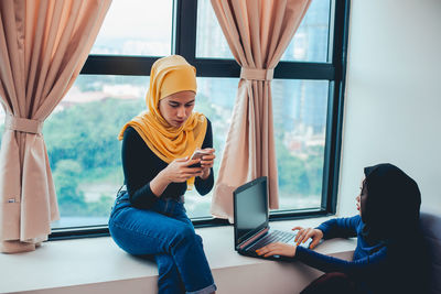 Young woman using mobile phone while sitting on window