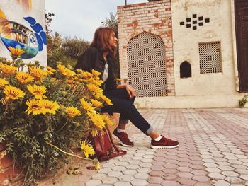 Side view of young woman sitting on footpath against buildings in city