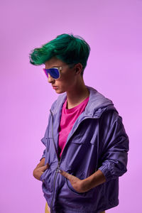 Side view of stylish female with short green dyed hair in sunglasses with blue lens standing against purple in studio