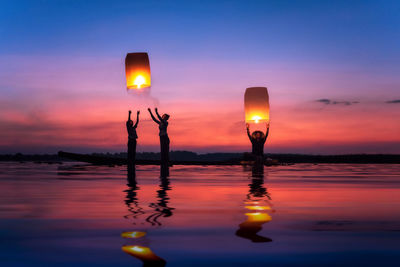 Silhouette family releasing illuminated lanterns while standing in sea at dusk