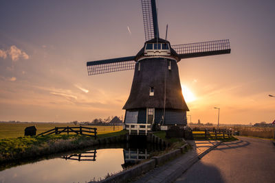 Traditional windmill by canal against sky during sunset