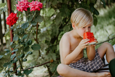 Shirtless boy eating watermelon while sitting against plants