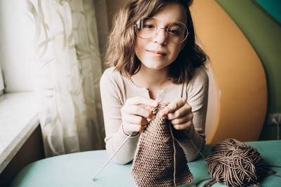 Teenage girl in a beige sweater, wearing glasses knitting needles standing by the window. 